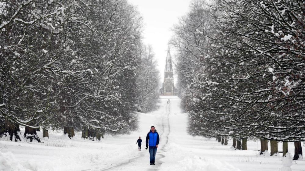 A man walking in the snow in Studley Royal park in North Yorkshire. He is on a path with bare-branched trees on either side. Behind him, at the end of the path, the spire of a church can be seen. 