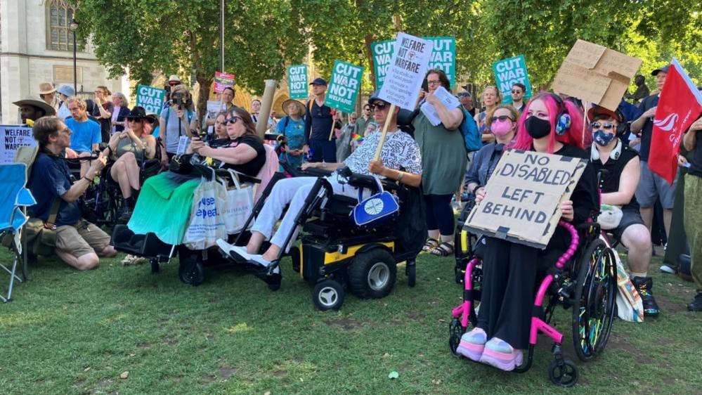 A group of people, some standing and some sat in wheelchairs, are gathered in a park. They are carrying banners that say things including 'cut war, not welfare'.