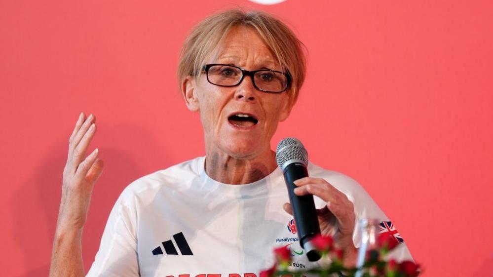 A woman with short blonde hair and glasses, pictured in a white t-shirt with the Paralympics GB logo on the right and the Adidas logo on the left. She is wearing glasses and speaking into a microphone that she is holding in her left hand. She is pictured against a light red background. 