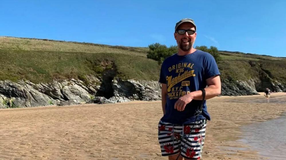 The photo shows Gavin stood on a beach with a grassy cliff in the background. He smiles at the camera while wearing a blue t-shirt with yellow writing and white and blue stripped shorts. He also wears sunglasses and a baseball style cap.