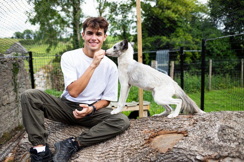 Kyle Thomas pictured in a white tshirt, green cargo pants and black trainers, feeding a white dog, sitting on tree bark with greenery and fences in the background