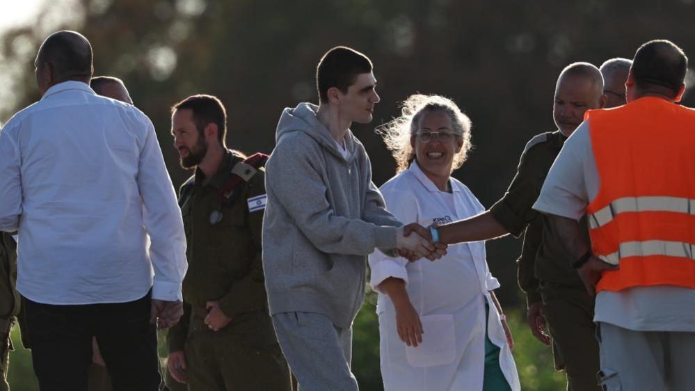 Maxim Herkin, wearing a grey, jumpsuit shakes hands with an Israeli officer as a medic in a white overcoat looks at him smiling