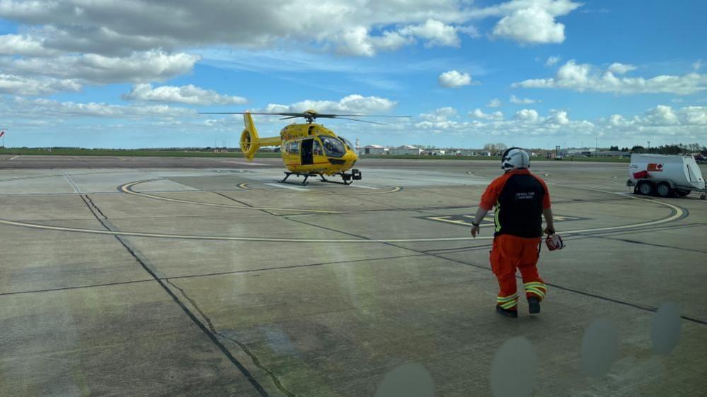 Helicopter standing on airport runway with person in orange outfit in the foreground