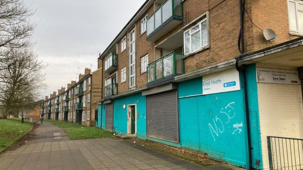 Part of Kings Heath with a unit boarded up, with blue painted wood in the foreground. A broken sign says "Kings Heath Centre". There are flats above the unit and continuing into the distance. There is a slabbed pavement running past the flats and an area of green to the left, with trees.