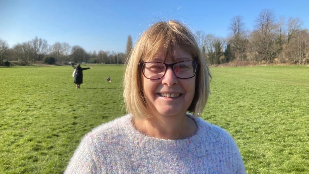 Wendy Randall with medium-length light brown hair and glasses, wearing a light-coloured sweater and standing in a grass field with trees behind. A woman is visible in the background waving her arms as she walks a dog.