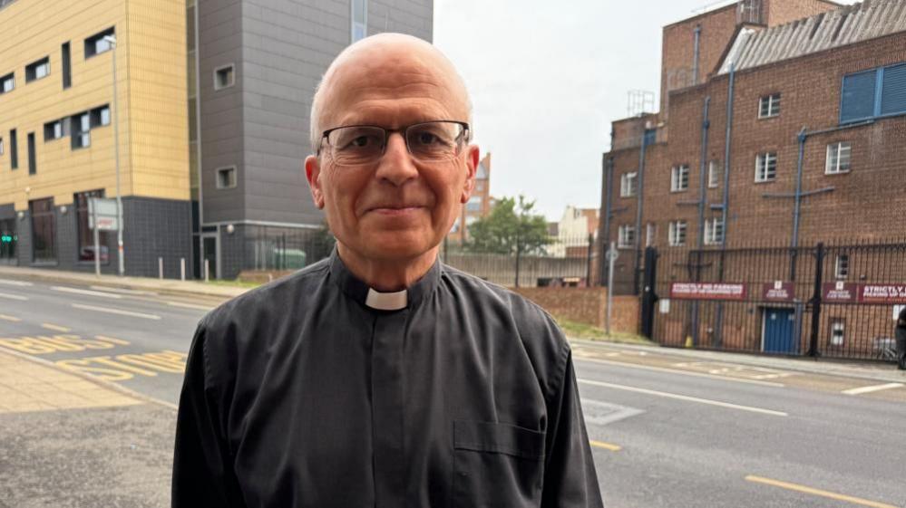 George Sarmezey in a black shirt with a white dog collar stands outside by a road smiling at the camera.