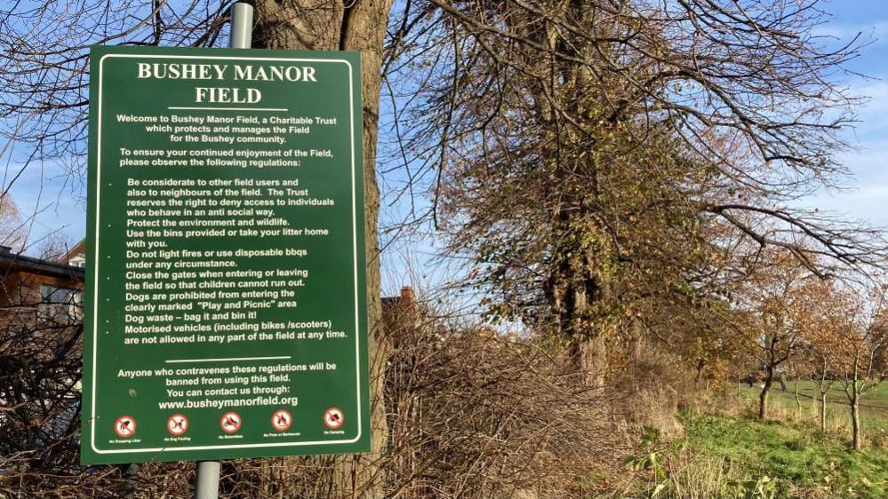 A green sign saying BUSHY MANOR FIELD. The sign lists a number of regulations people are asked to observe. In the background is a hedge, trees and grass 