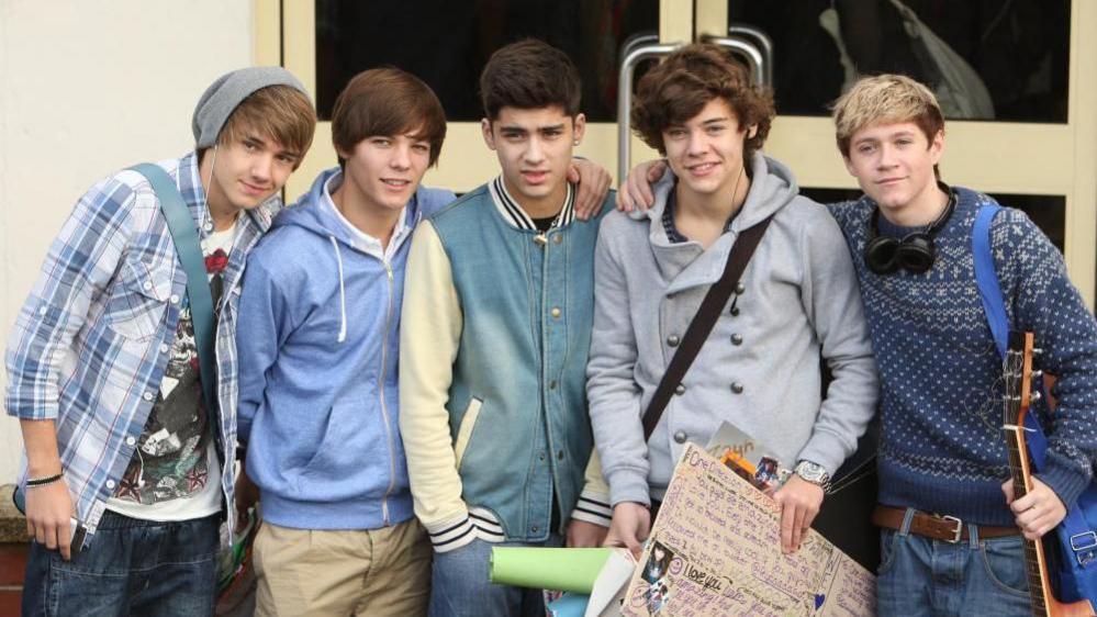 All five members of one Direction looking boyish in a picture from 2010, in which Harry Styles holds a sign written by a fan while a young Louis Tomlinson holds a guitar.