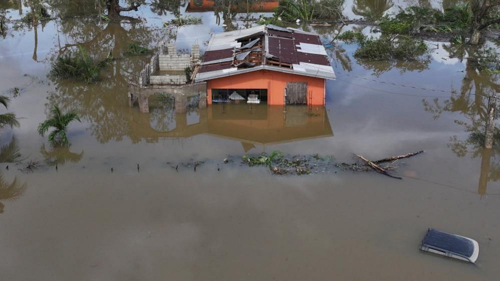 An orange house with a partially collapsed roof and broken solar panels is surrounded by muddy brown floodwater that reaches halfway up its walls. Debris, including fallen branches and wooden planks, floats nearby. A silver car is almost completely submerged in the water to the right of the house. The surrounding area shows palm trees bent or stripped of leaves.