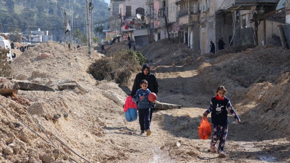 Two children and their mother walk along a rubble filled street carrying bags and a football in Tulkarm camp