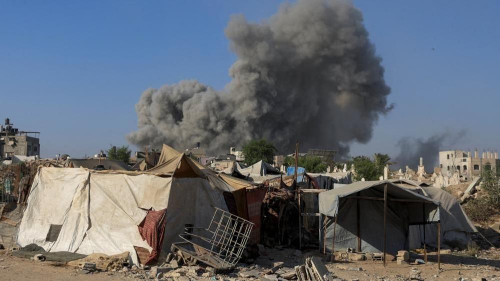 Ramshackle tent-like structures, one has a kind of a metal grid in front, with a large plume of smoke behind them and two half-finished barely-standing building on either side, in Gaza City on 24 August 2025