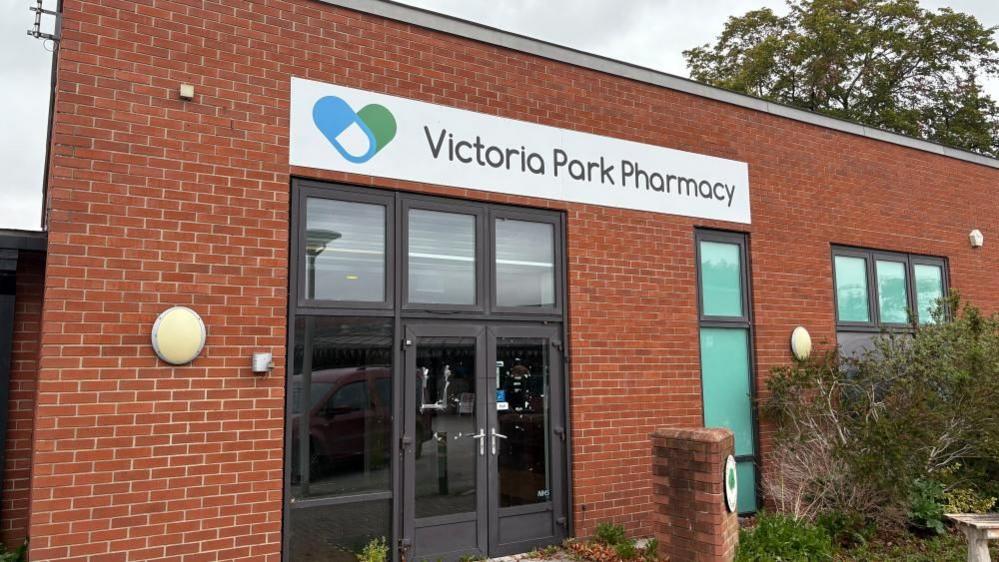 The photo shows a rectangular, one storey red brick building. A large sign above a double glass door reads "Victoria Park Pharmacy". No light comes from inside, with an overcast sky above.