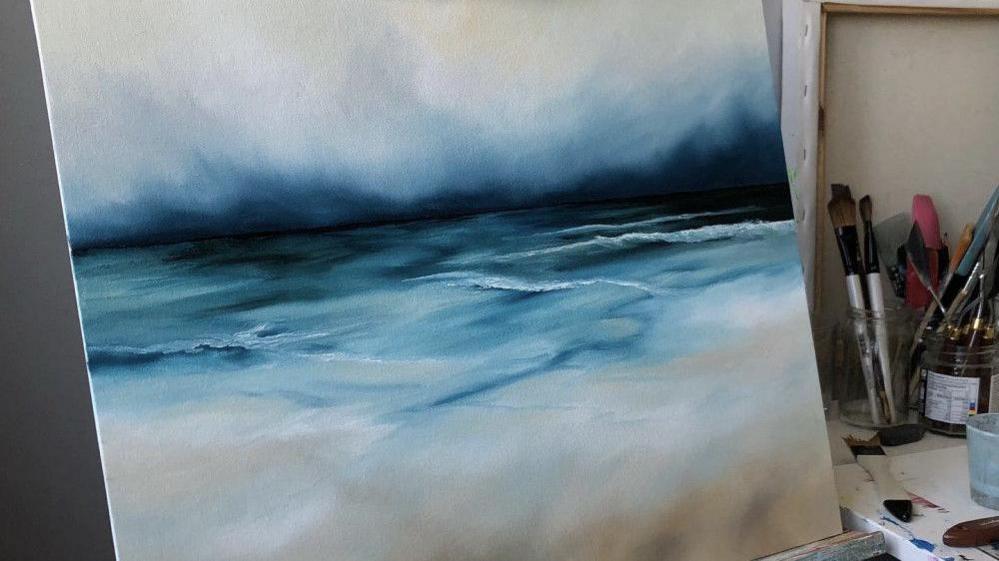 A stormy picture of the sea and shore painted on a square canvas, propped up on an easel. There are dark blues on the horizon, and browns on the bottom for the sand. There is a pot of paintbrushes on the table on the right