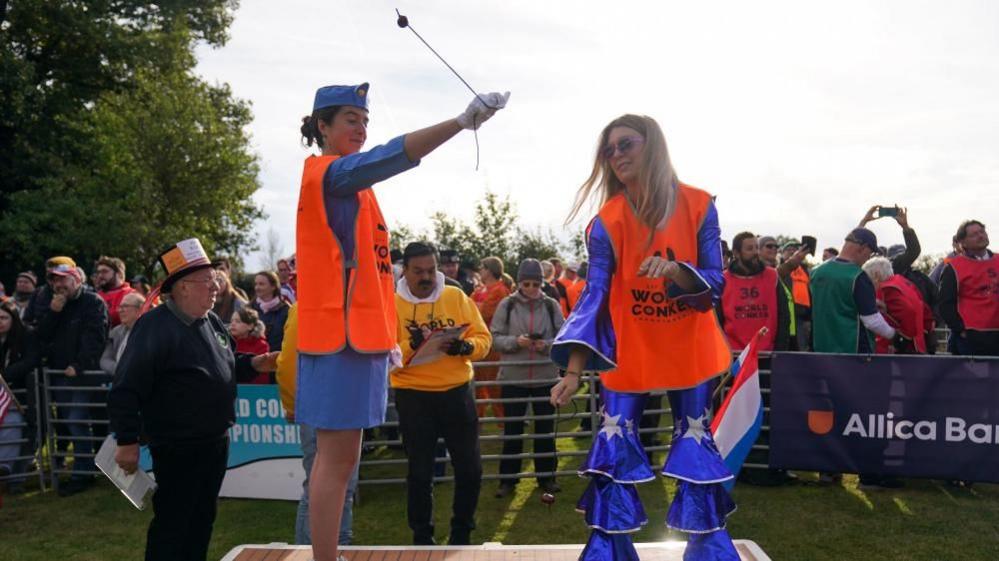 Two female competitors, both in blue fancy dress outfits, on a platform taking part in the competition while being watched by judges and spectators