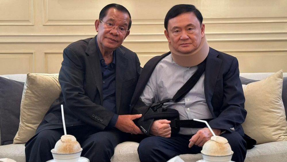 Hun Sen (l) holds the arm of Thaksin Shinawatra (R) as they sit on a grey sofa with beige and grey cushions. They each have a coconut with a straw in front of them served in a blue and white ceramic bowl on a blue and white saucer.