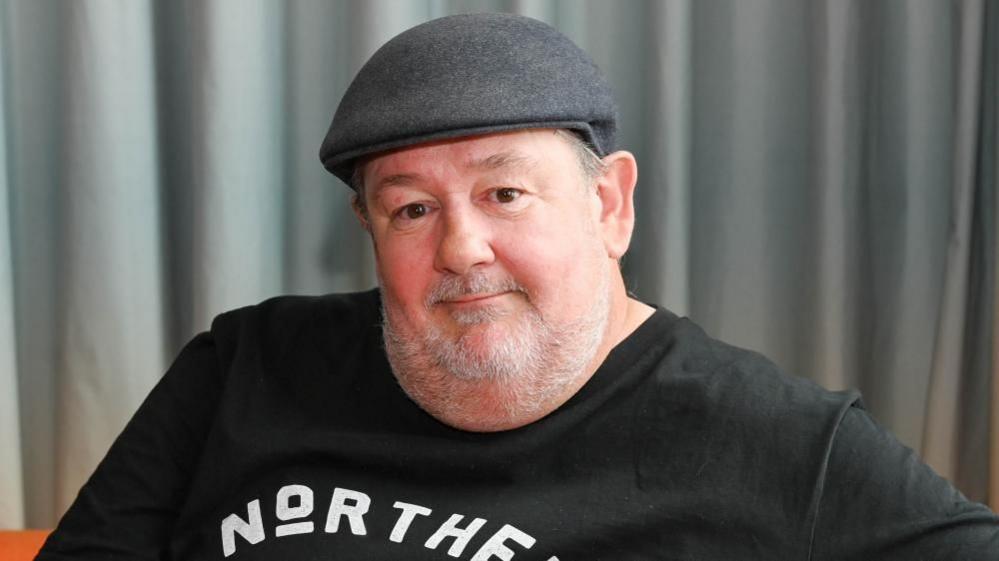 Johnny Vegas wearing a dark flat cap and a black T-shirt with the word “NORTHERN” printed in bold white letters across the chest. The background consists of grey curtains.