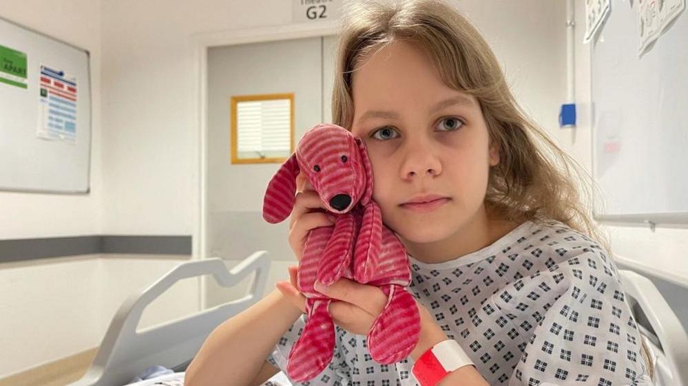 A girl looks nervously into the camera. She is wearing in a hospital gown and sits in a hospital bed. She is cuddling a little pink dog.