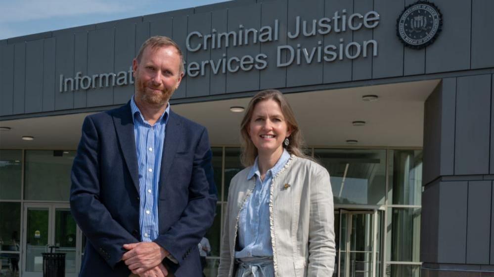 A man and a woman stand in business wear in front of a grey, modern building. The front on the building reads 'Criminal Justice Information Services Division' next to the FBI logo. 