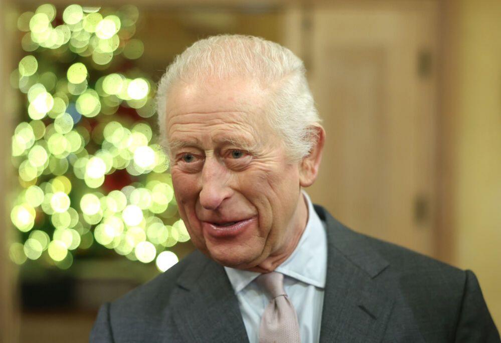 King Charles III smiles at a Christmas event at Highgrove Gardens. He has white hair and wears a dark grey suit with a pink tie and Christmas lights can be seen in the background