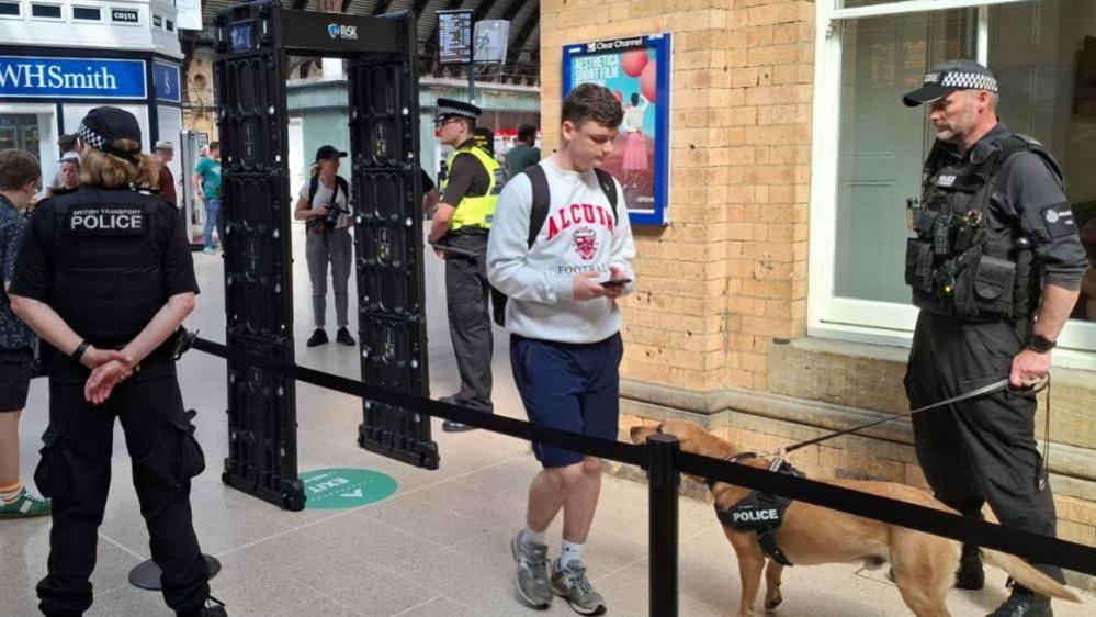 A young man walks past a police dog with a knife arch metal detector behind him. Other police officers are visible patrolling the area.