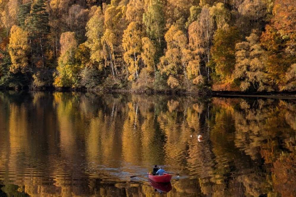 Trees with orange and green leaves reflecting onto Loch Faskally. There is two people in a kayak in the foreground.