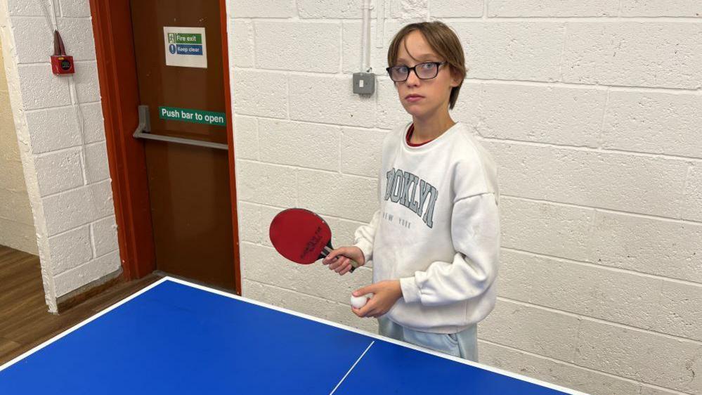 Carmen is standing at a table-tennis table. She is holding a red bat in her right hand and a ball in her left hand. She is wearing spectacles and a white jumper.