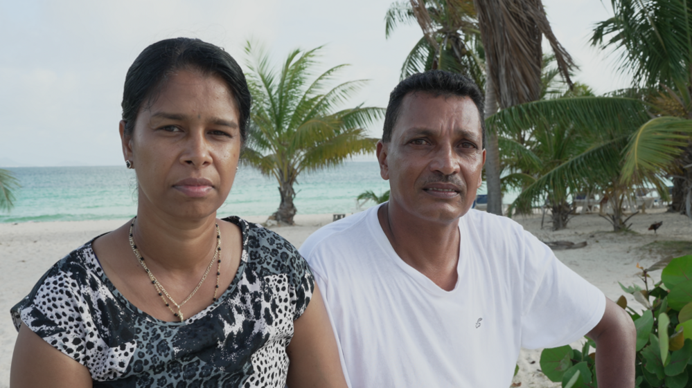 Mr and Mrs Seesahai stare at the camera with solemn faces; they are on a beach in Anguilla, with palm trees and the sea visible in the background