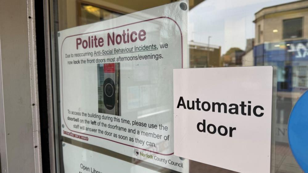 Notices in the door of Gorleston Library bear the text: "Automatic door" and another: "Polite notice; Due to recurring antisocial behaviour incidents we now lock the front doors most afternoons/evenings. To access the building during this time, please use the doorbell on the left of the door frame and a member of staff will answer the door as soon as they can."