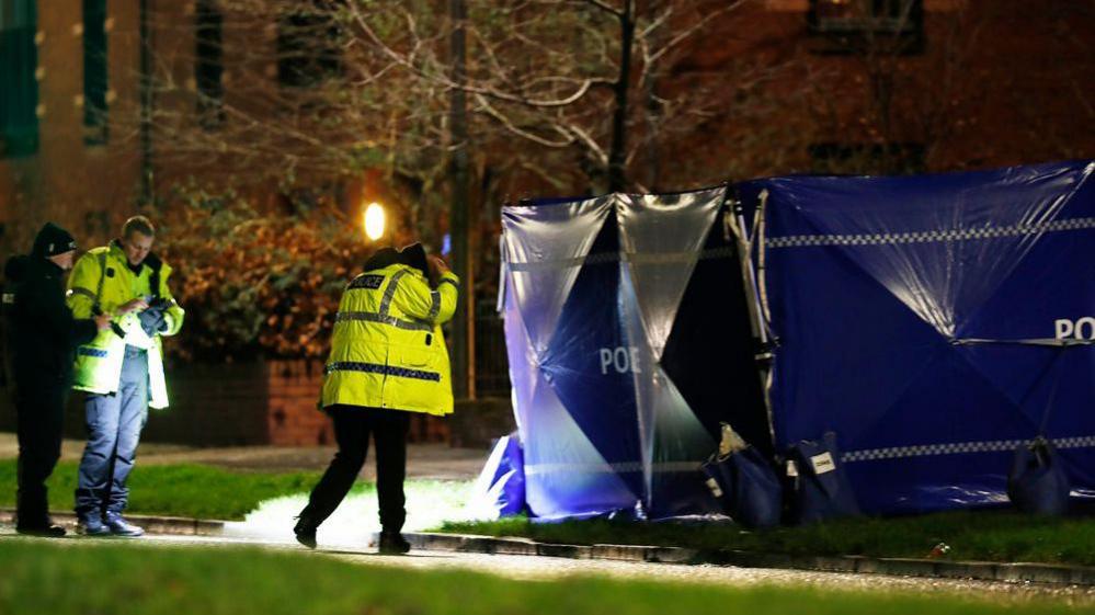 Police stand with torches outside a blue police tent erected on a pavement.