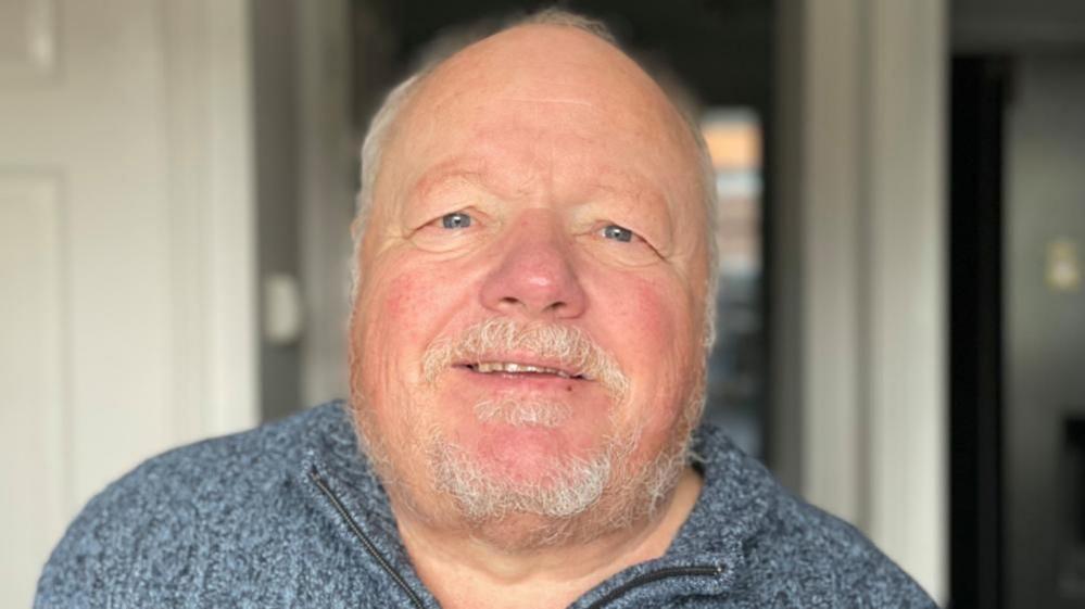 Mr Haskell is wearing a blue zip fleece. He is standing in the hallway looking directly at the camera in front of the white walls and doorway of his home.