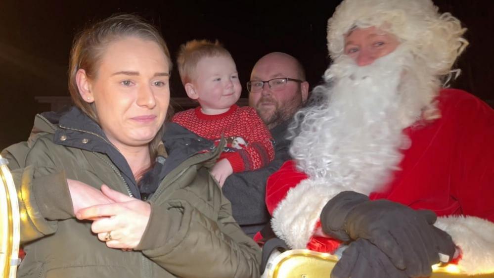 A couple holding a toddler, with a man dressed as Santa