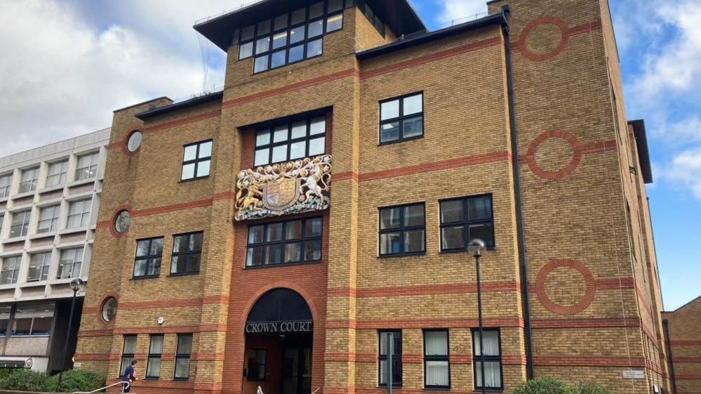 The outside of St Albans Court Court, showing a brick building, with a courts of justice coat of arms. with black windows, a large entrance and steps up to the building. 