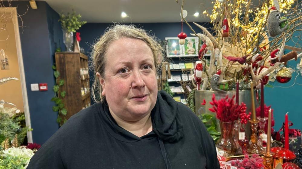 Gill Cowper stood in a florist with Christmas displays behind her. She is wearing a black hoodie and looking at the camera