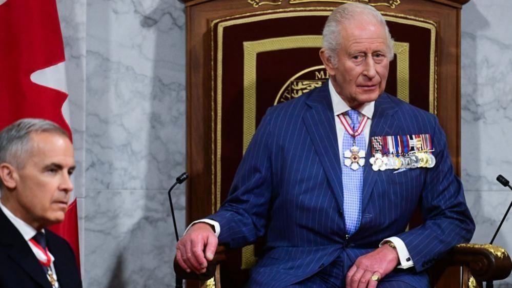 Canada Prime Minister Mark Carney and King Charles III on the Senate floor during the State Opening of the Parliament of Canada, in the Senate Chamber.  Both have neutral expressions. The King sits on the Throne and wears a blue pinstripe suit and a number of medals. 