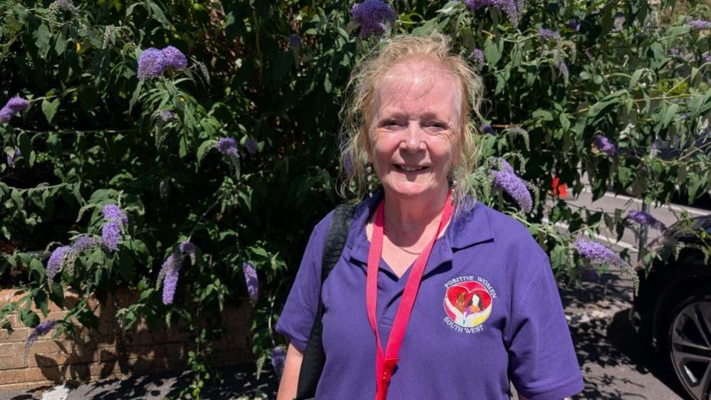A headshot of Positive Women South West founder Bonnie Holt. She is wearing a purple polo shirt and red lanyard. She is standing in front of a bush of purple flowers.