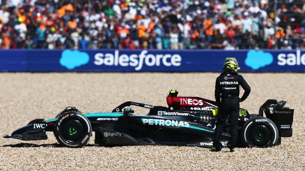 Lewis Hamilton stands with his hand on his hip looking at his Mercedes beached in the gravel trap