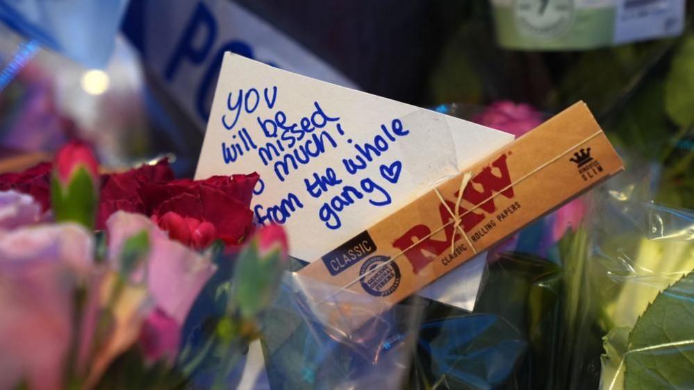 A bunch of flowers with a message that reads: "You will be missed so much! From the whole gang." A packet of cigarette rolling papers is stuck to the message.