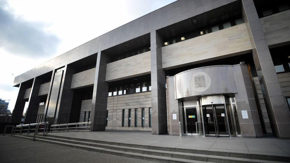 A general view of Glasgow Sheriff Court, a grey building with a grey exterior and a entrance with a revolving door which has a crest above it. 