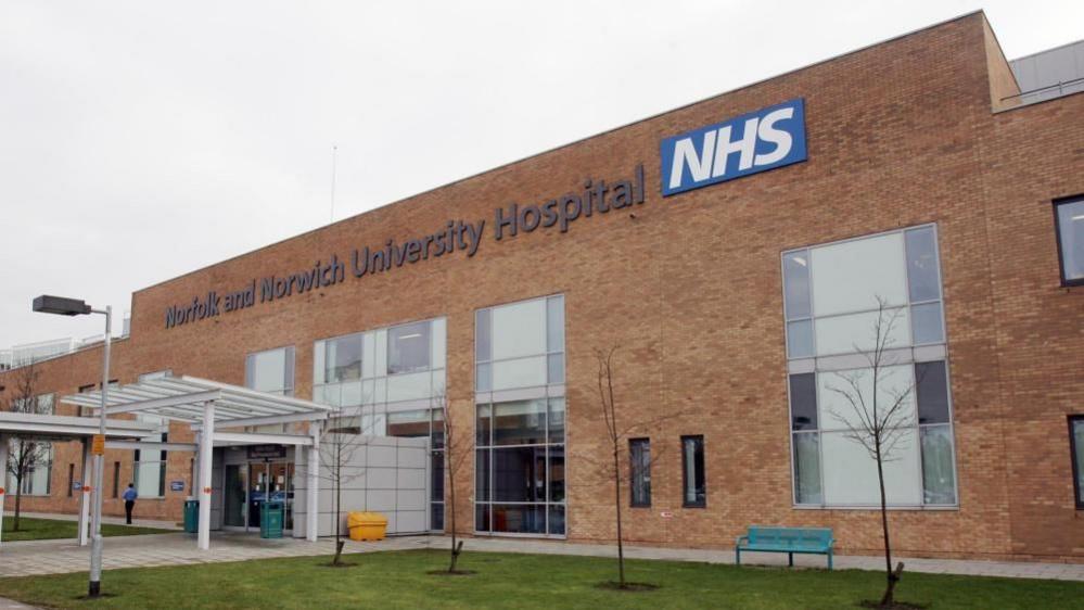 The Norfolk and Norwich University Hospital, bearing NHS signage stating its name, is a large yellow brick building, featuring large windows, and canopies, and lawns to the front.