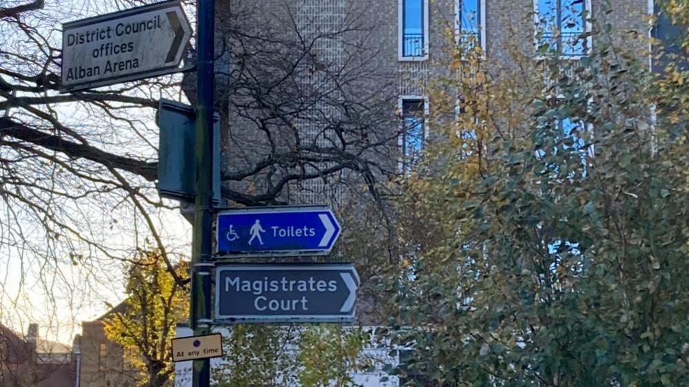 Three signs attached to a sign post in front of trees and a building. A white sign says "District Council offices Alban Arena; a blue sign has logos of a man walking an a wheelchair and says "Toilets"; a second blue sign says "Magistrates Court".