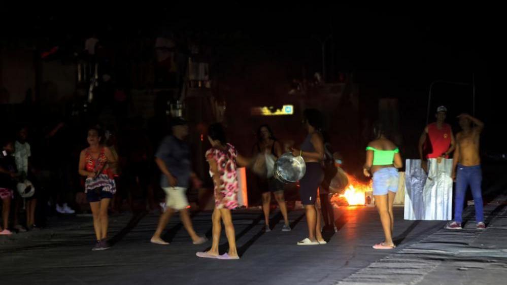 People protest against power outages in Havana on 7 July 2026. One person can be seen holding a large pot while another is banging a metal sheet. In the background burning rubbish can be seen on the street. 