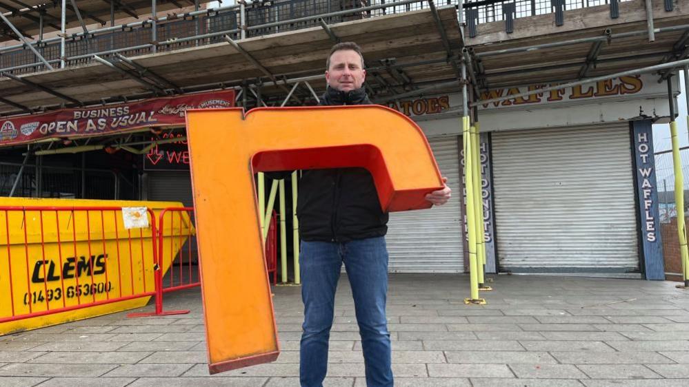 Joseph Abbott, wearing blue jeans and a black jacket, stands outside the Britannia Pier, holding a letter 'R' made from orange acrylic, which once was mounted on the pier façade. 