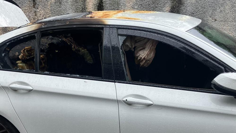 The white car displays broken windows and substantial burns inside and on the roof.