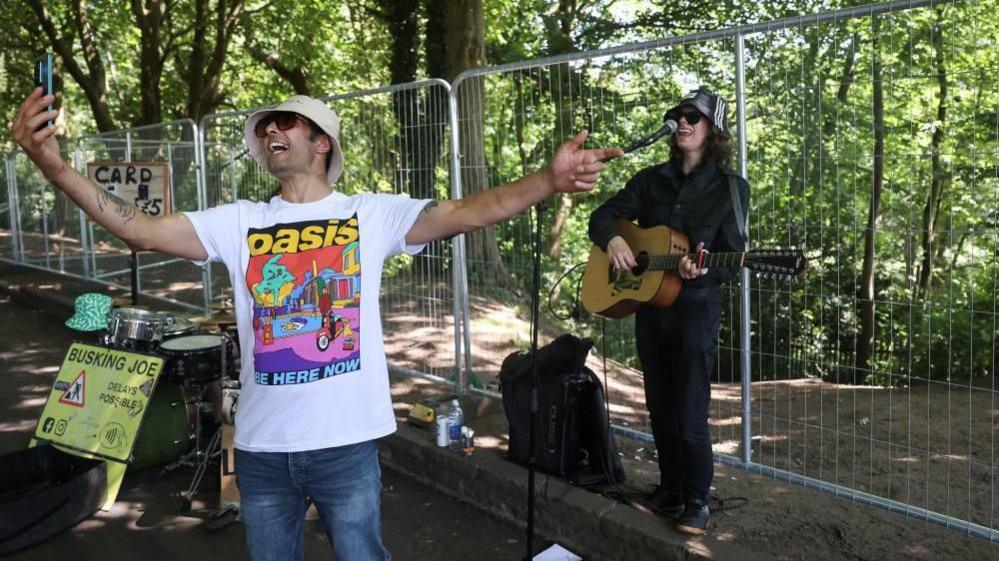 A man wearing an Oasis T-shirt and white bucket hat is singing while taking a selfie with his phone with a busker playing guitar behind him, in front of railings. A mock road sign says 'Busking Joe' and 'delays possible'. The busker is also wearing a bucket hat.