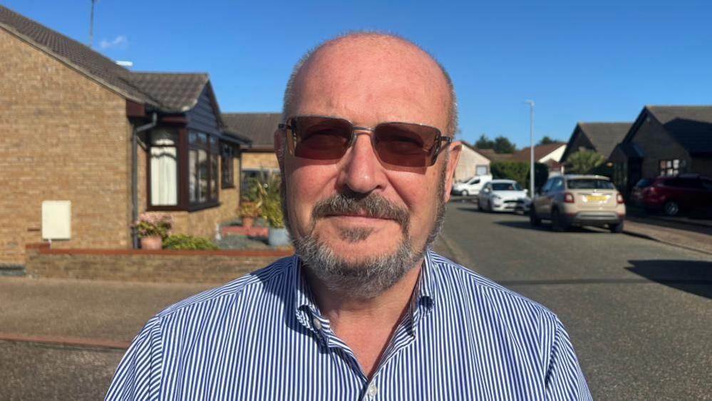 County councillor Graham Plant, wearing spectacles. He has close cropped greying hair and beard with moustache. He is wearing a blue and white vertical-striped shirt and is standing in a street of bungalows and parked cars