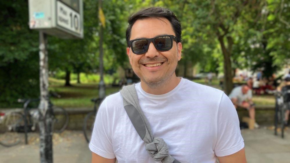 A man wearing a white T-shirt and sunglasses smiles at the camera. He has a grey jumper over his shoulder. Trees are in the background, with people sitting on benches too.