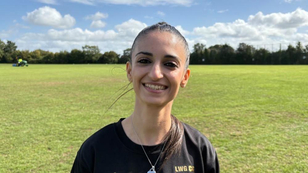 Kiki Christofi with long dark hair tied back. She is wearing a black LWSCC top and necklace with large pendant. There is a cricket pitch behind and hedges beyond.