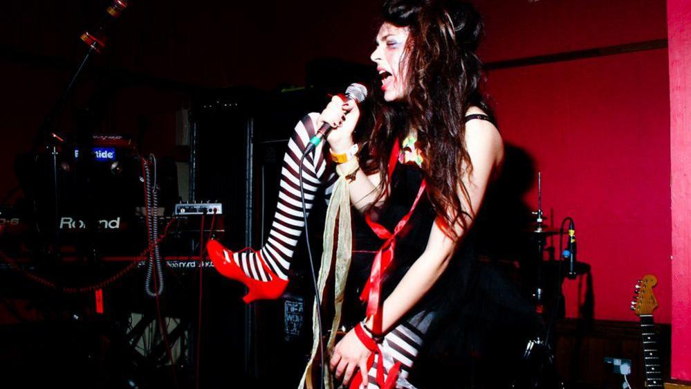 A woman singing into a microphone with a long red ribbon around her and stripy black and white tights, her hair is dishevelled and her make-up is smudged.