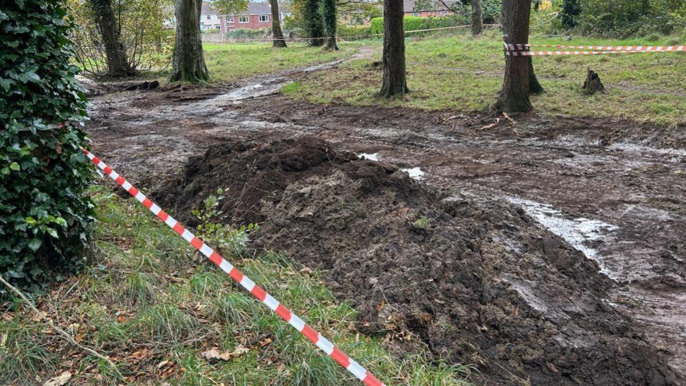 Sewage and mud is piled up alongside a path of trees. A red and white tape has been placed to keep the public away from affected areas.