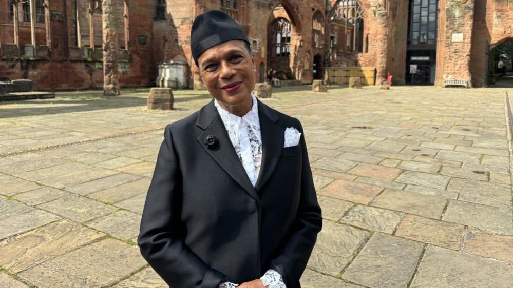 The lead singer of The Selecter standing outside Coventry Cathedral. She is wearing a black jacket and trademark black hat with a white lace shirt